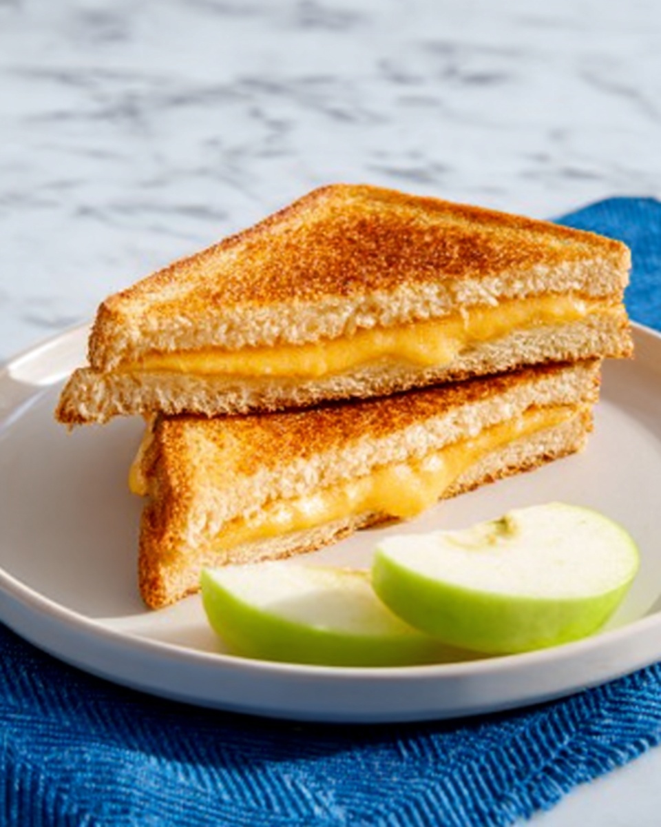 Sándwich de Queso y Champiñones Recipe 4 The image shows two triangular grilled cheese sandwiches stacked on a white plate, each sandwich displaying golden-brown toasted bread with melted yellow cheese between the layers. The sandwiches are positioned on the left side of the plate, while two green apple slices with smooth skin and a fresh texture rest neatly on the right side. The plate sits on a white marbled surface with a blue cloth underneath it. The lighting highlights the crispy texture of the bread and the soft melted cheese photo taken with an iphone --ar 4:5 --v 7
