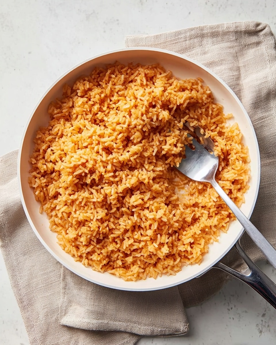 A white frying pan filled with cooked rice that has an orange color, showing a texture of separate grains slightly sticking together. A silver fork is resting inside the pan, partially mixed into the rice on the right side. The pan sits on a folded beige cloth napkin, all placed on a white marbled surface. The lighting is soft, highlighting the warm tones of the rice. photo taken with an iphone --ar 4:5 --v 7