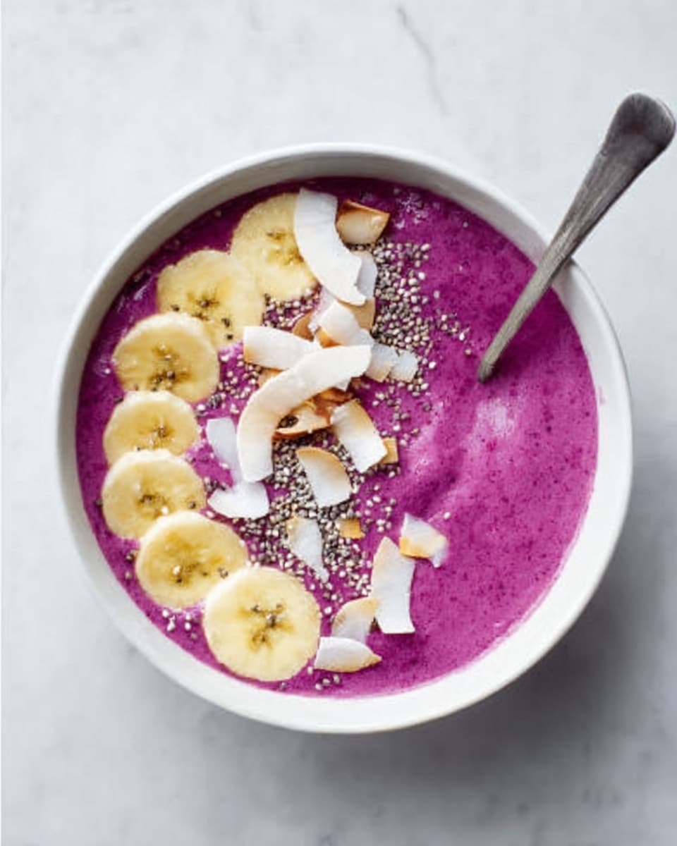 A white bowl sits on a white marbled surface, filled with a smooth, bright purple smoothie base. On top, there are thin slices of yellow banana lined up on one side, scattered small white seeds with tiny black specks in the middle, and a few larger pieces of white and light brown coconut flakes spread around. A silver spoon rests inside the bowl, dipping into the vibrant smoothie. Photo taken with an iphone --ar 4:5 --v 7