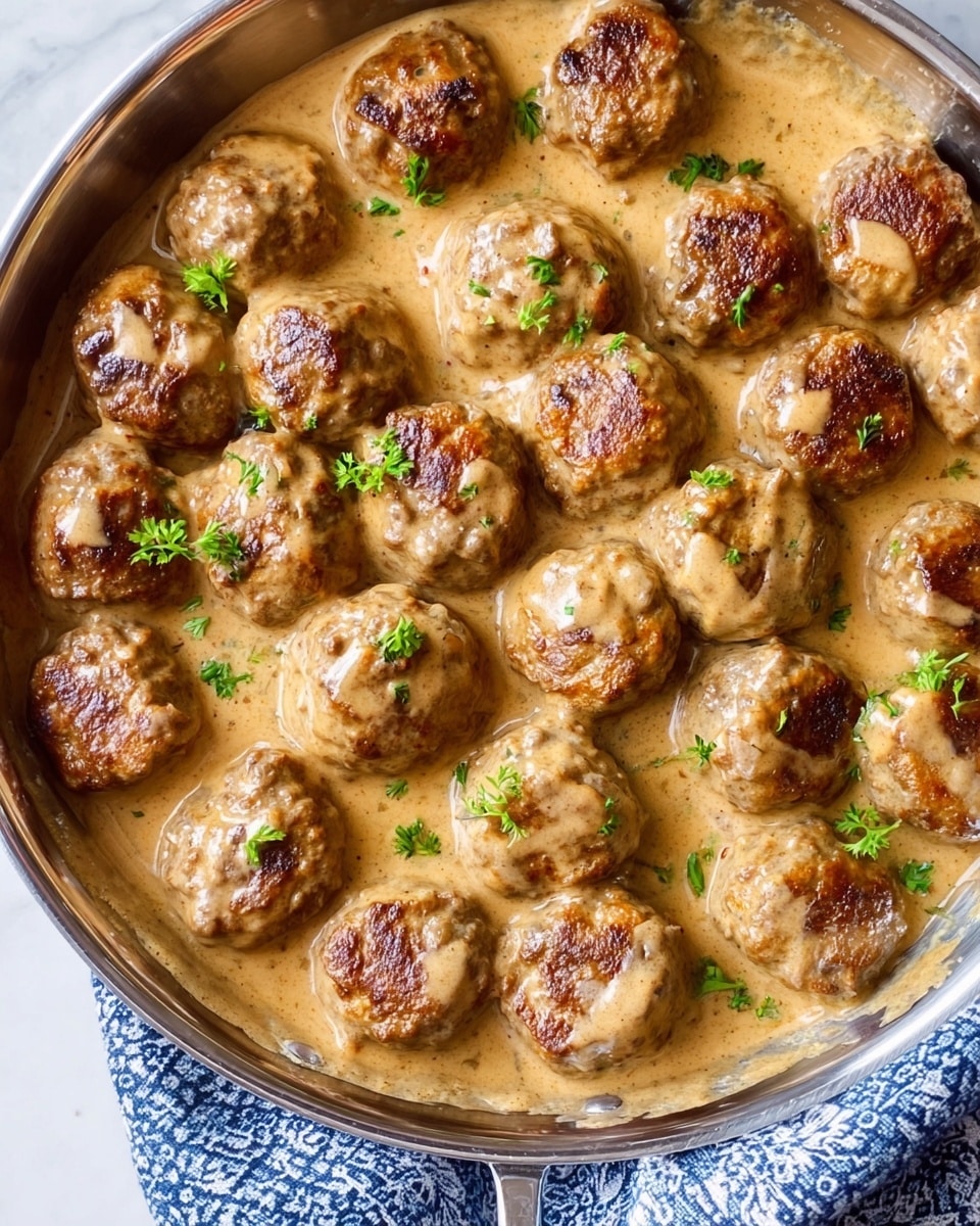 The image shows a round stainless steel pan filled with small, round meatballs covered in a light brown creamy sauce. The meatballs are arranged in close rows, and each one has a slightly rough texture with some small green herbs sprinkled on top, adding a fresh splash of color. The creamy sauce looks smooth and coats the meatballs evenly. The pan sits on a white marbled surface with a blue and white patterned cloth partially visible under the pan’s edge. Photo taken with an iphone --ar 4:5 --v 7