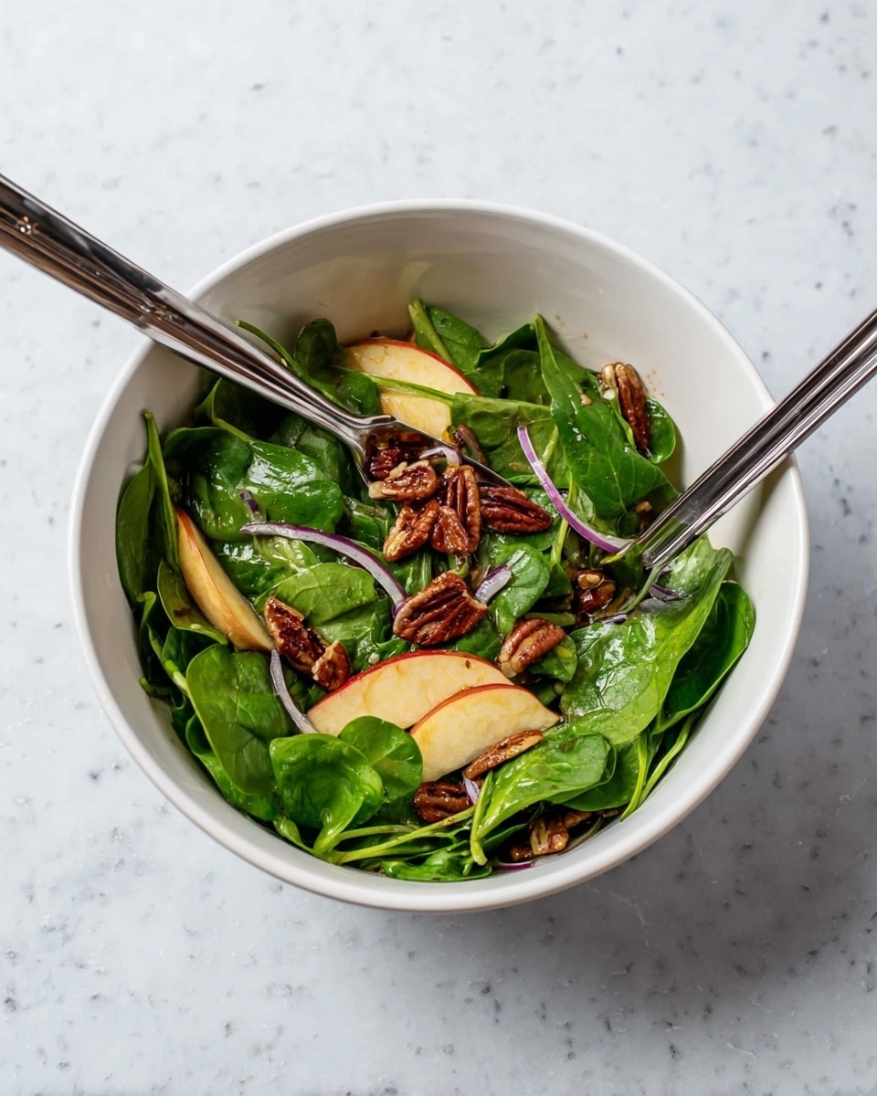The image shows a white bowl filled with a fresh salad sitting on a white marbled surface. The salad has three main layers: at the bottom, there are bright green spinach leaves with a smooth texture; on top of the spinach are thin, curved slices of apple with pale yellow flesh and red skin edges; scattered around and above the apple slices are shiny, dark brown pecan nuts and thin, purplish-red slices of onion rings. Two silver metal salad tongs with black silicone tips rest inside the bowl on opposite sides, partially submerged in the salad. The salad looks fresh and lightly mixed. Photo taken with an iphone --ar 4:5 --v 7