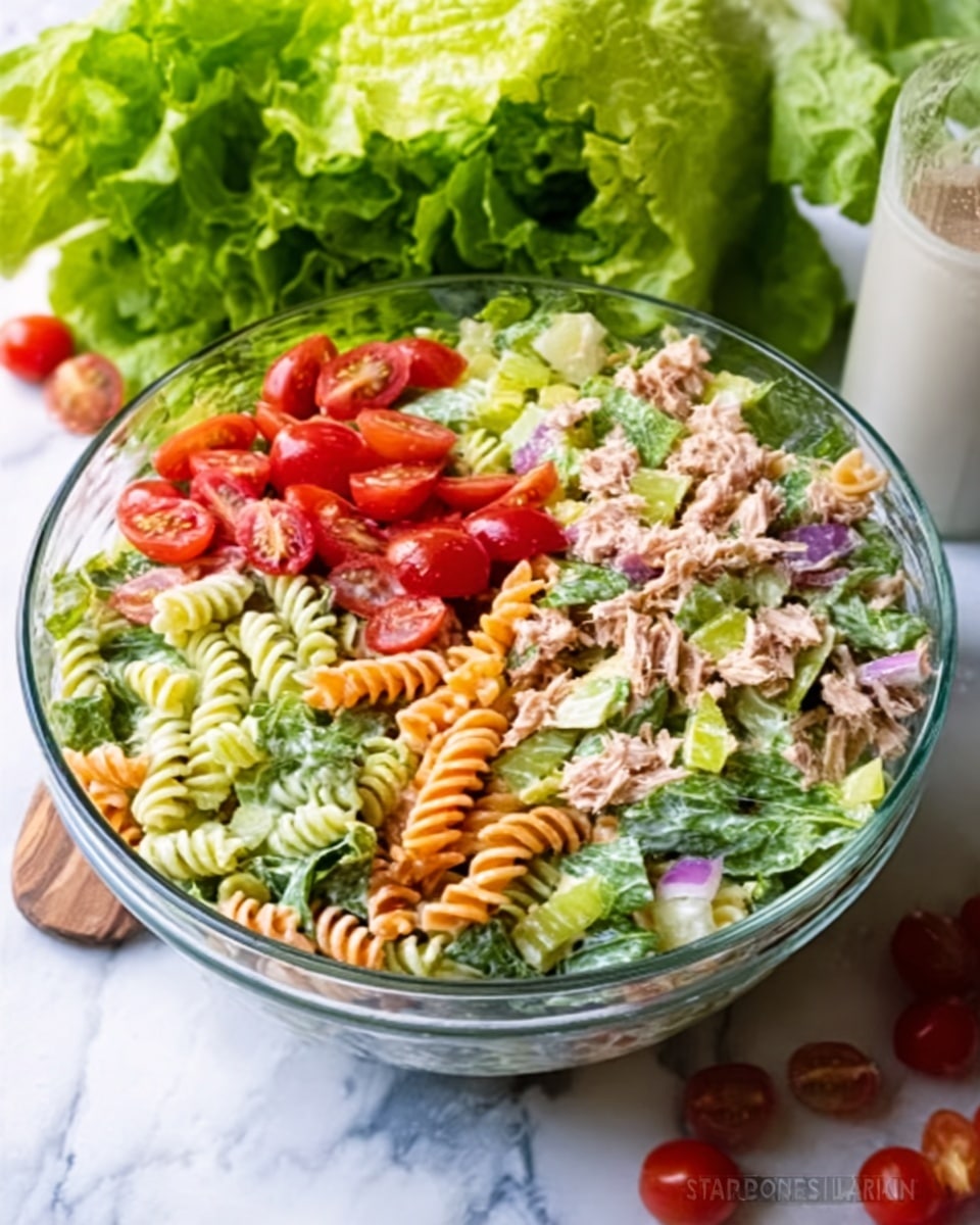 A clear glass bowl filled with a colorful rotini pasta salad on a white marbled surface. The salad contains curly rotini pasta, fresh arugula, chopped artichoke hearts, halved cherry tomatoes, and grated Parmesan. Behind the bowl, there is a head of fresh arugula. The scene is bright with natural light.