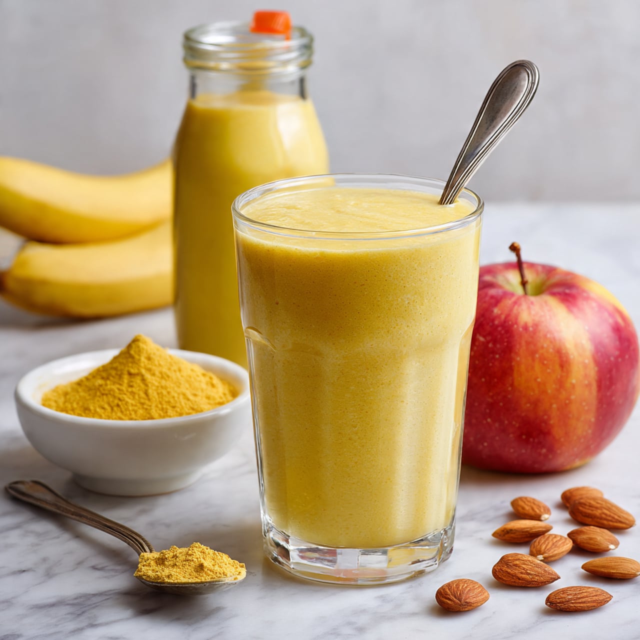 A clear glass filled with thick, smooth yellow smoothie placed on a white marbled surface, with a silver spoon inside it. Behind the glass, there is a transparent bottle filled with the same yellow liquid. To the left, a white bowl with a small amount of yellow powder sits beside a silver spoon holding some of that powder. Next to the bowl, a ripe banana lays flat. On the right side, an apple with a reddish-yellow skin is placed next to a small glass jar containing more of the yellow powder and sealed with an orange clip. A few scattered almonds are spread near the glass. Photo taken with an iphone --ar 4:5 --v 7
