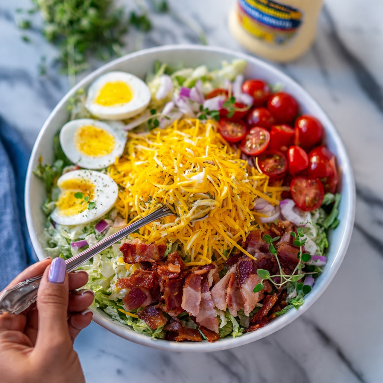 A white bowl filled with a colorful salad sits on a white marbled surface. The salad has many layers: the bottom layer is shredded green lettuce, topped with bright yellow shredded cheese. Scattered over the cheese are red cherry tomato halves, slices of white boiled eggs, and pieces of cooked pink bacon. There are small green leafy herbs sprinkled on top for freshness, and a spoon is inside the bowl with a woman's hand holding it. In the background, a bottle of ranch dressing is partially visible. Photo taken with an iphone --ar 4:5 --v 7