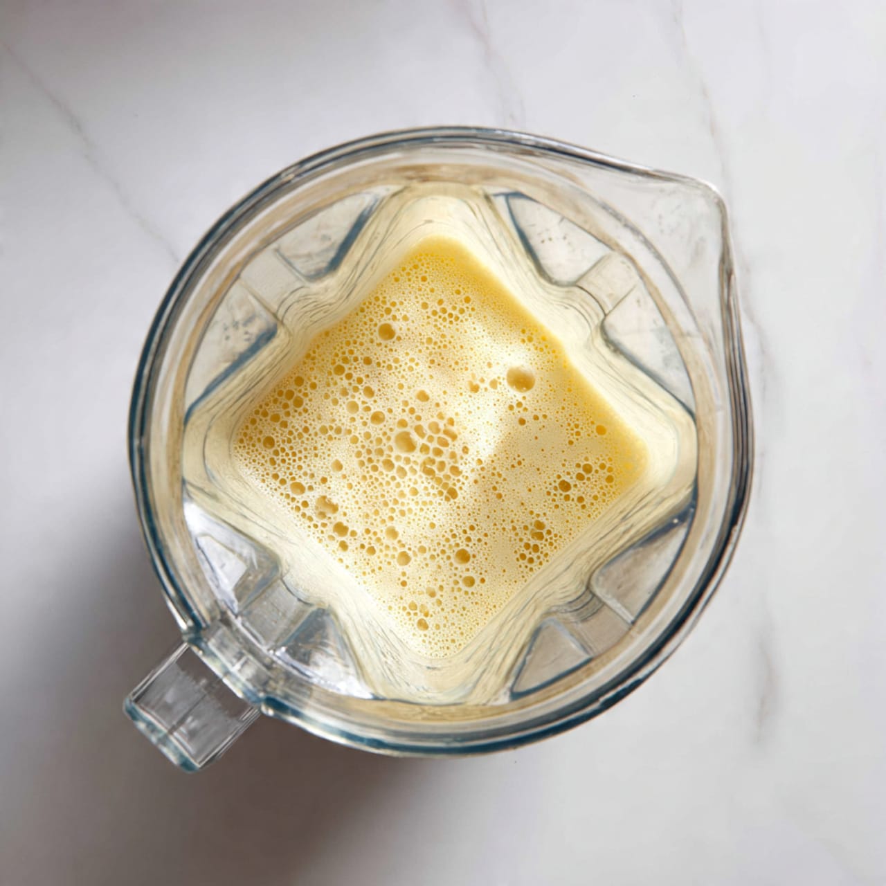 A clear blender container is shown from above, filled halfway with a light yellow bubbly liquid with many small bubbles forming a foamy texture on top. The blender has four triangular, raised shapes inside along the sides that catch the light, and the handle is positioned on the left side. It sits on a white marbled surface. photo taken with an iphone --ar 4:5 --v 7