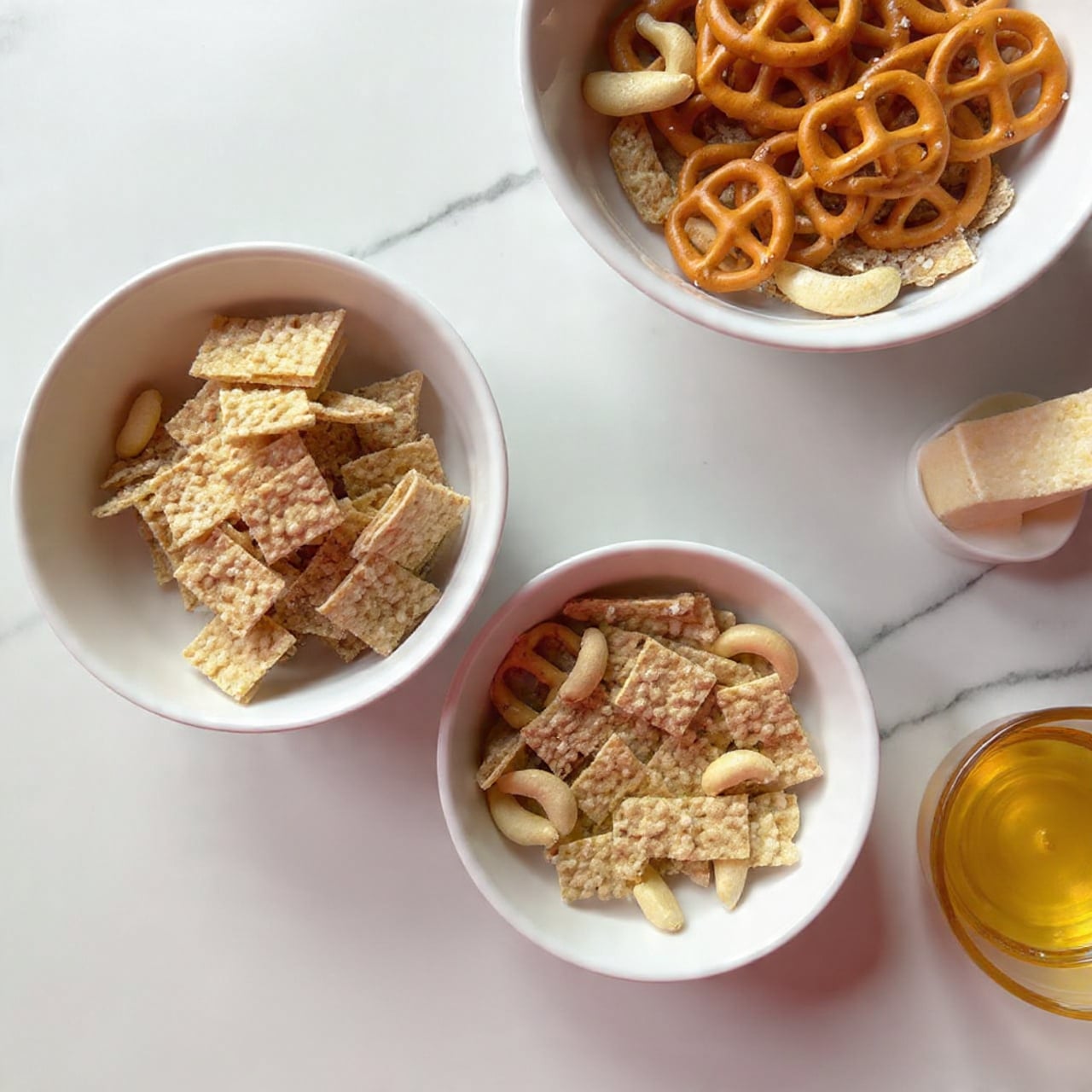 The image shows a white bowl filled with a mix of snack items on a white marbled surface. The bowl contains three visible layers: the bottom layer is made of light brown square cereal pieces with a textured pattern, the middle layer has several golden brown pretzels shaped like small twists, and the top layer shows scattered light beige cashews with smooth curved shapes. The snacks fill the bowl fully and look crispy. Next to this bowl, a part of another white bowl filled with the same mixture is slightly shown. Photo taken with an iphone --ar 4:5 --v 7