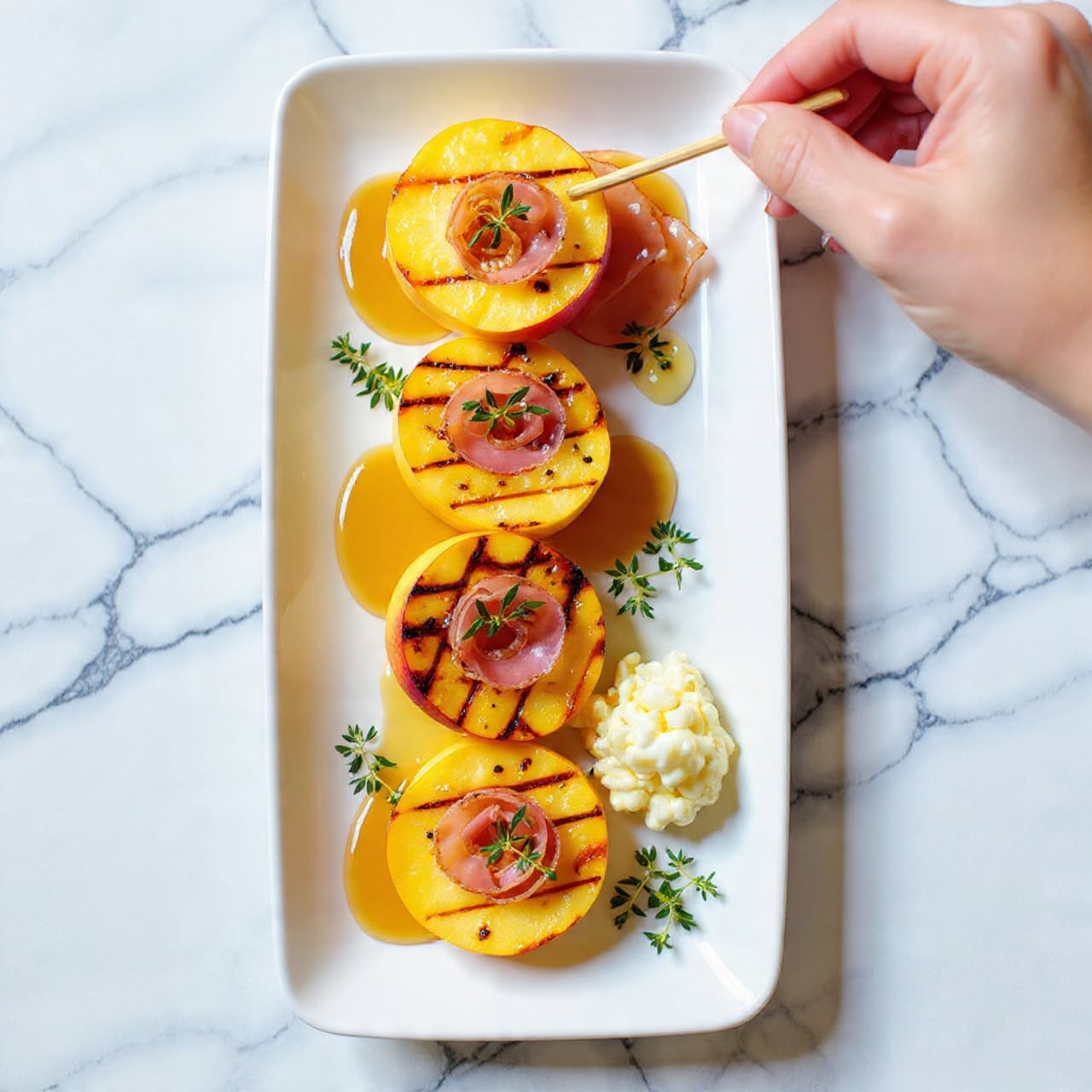 The image shows a long white rectangular plate on a white marbled surface, holding several grilled peach halves arranged in a single row. Each peach half is golden with visible grill marks, slightly caramelized. On top of each peach half, there is a thin layer of crispy bacon with a shiny glaze, adding a reddish-brown color. Fresh chopped green herbs are sprinkled evenly over the peaches and bacon, adding a bright contrast. A light drizzle of golden syrup or honey glistens over the dish, enhancing the shine and texture. A woman's hand is using a wooden skewer to hold one of the peach halves. photo taken with an iphone --ar 4:5 --v 7