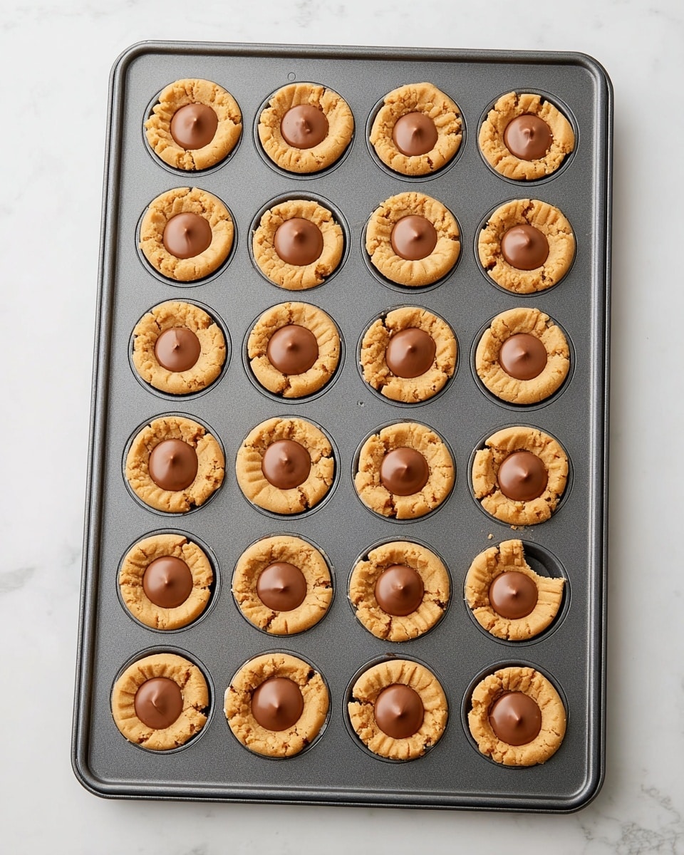 The image shows a dark gray baking tray with 24 round peanut butter cookies arranged in a 4 by 6 grid. Each cookie has a soft golden-brown color with slightly cracked tops, and sitting in the center of every cookie is a smooth, milk chocolate cup with a shiny finish. The cookies fill each round slot of the tray snugly, with one cookie on the bottom right having a small bite taken out, revealing a slightly crumbly texture inside. The tray is placed on a white marbled surface that gives a clean and bright background to the warm tones of the cookies and chocolate. photo taken with an iphone --ar 4:5 --v 7