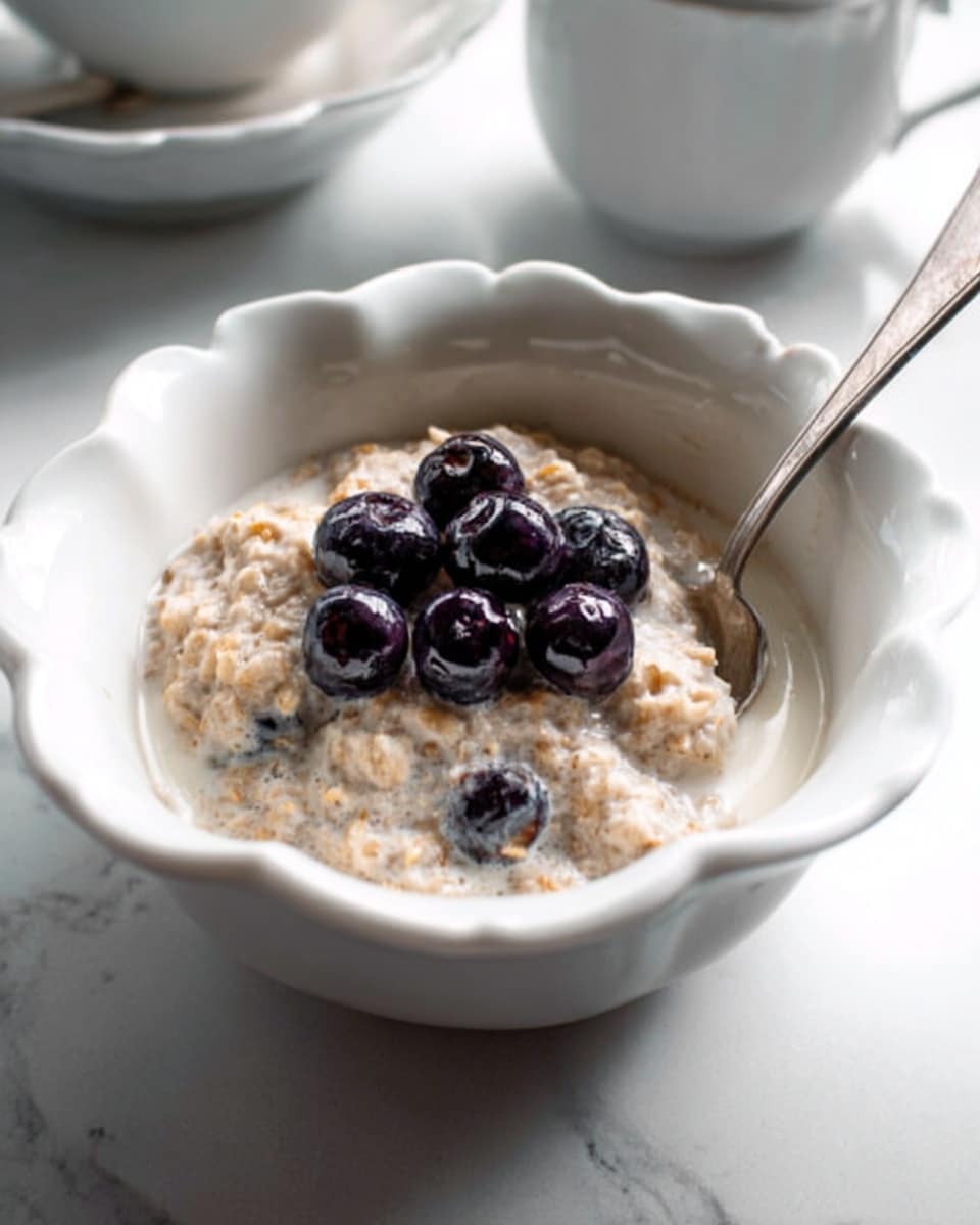 A white bowl with scalloped edges holds a creamy, light brown oatmeal soaked in milk, topped with a small pile of dark, glossy blueberries placed in the center. A silver spoon rests inside the bowl on the right side, partially submerged in the oats and milk. The bowl sits on a white marbled surface, with another white bowl and a white cup partially visible in the blurred background. photo taken with an iphone --ar 4:5 --v 7