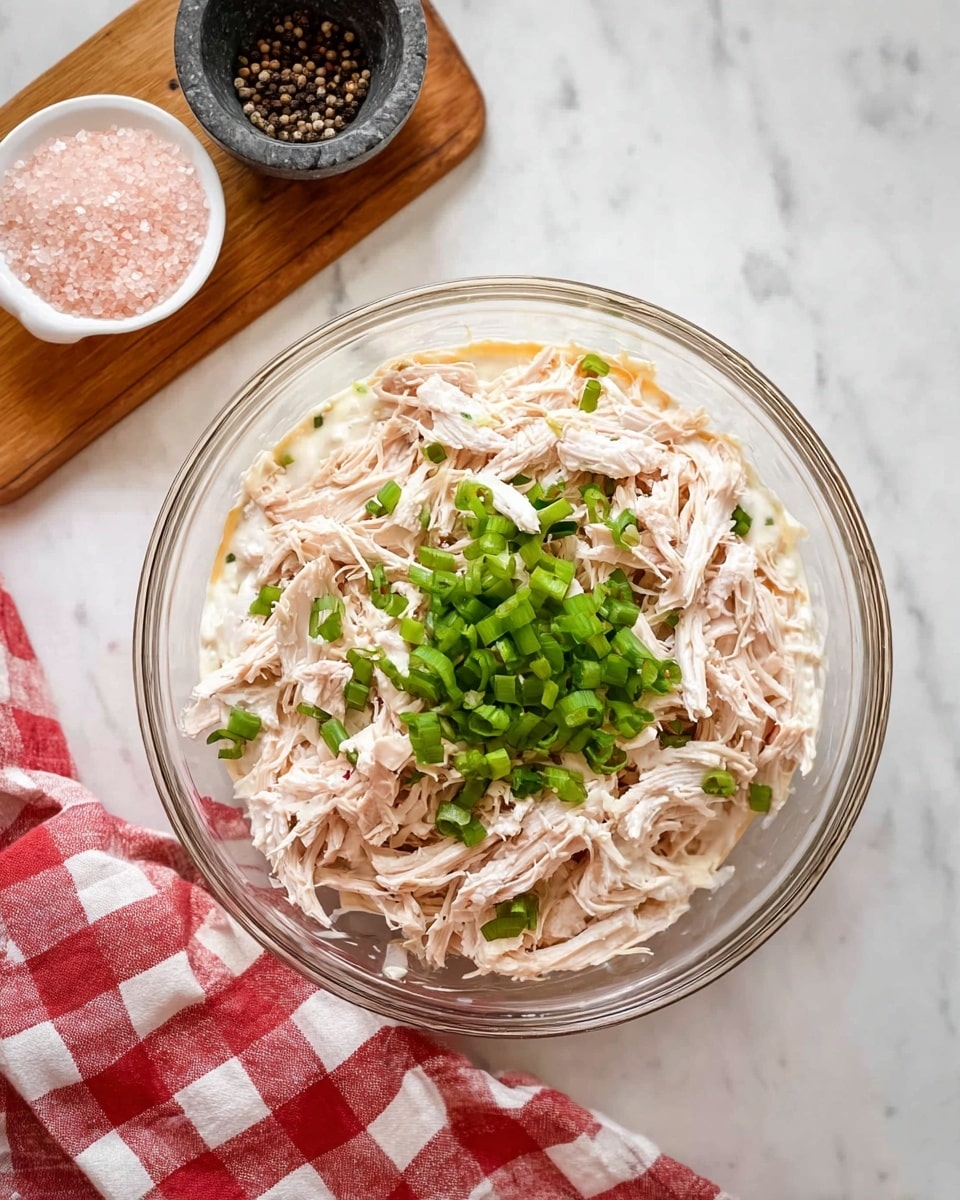 A clear glass bowl sits on a white marbled surface with a red and white checkered cloth partially underneath. Inside the bowl is a layered mixture starting with a base of creamy white sauce, topped with shredded pale pink chicken pieces spread evenly, and garnished in the center with chopped bright green spring onions. To the upper left of the bowl, a small wooden board holds a white container filled with pink salt and a small dark stone mortar and pestle with black peppercorns. The scene is bright and clean with a fresh kitchen feel, photo taken with an iphone --ar 4:5 --v 7