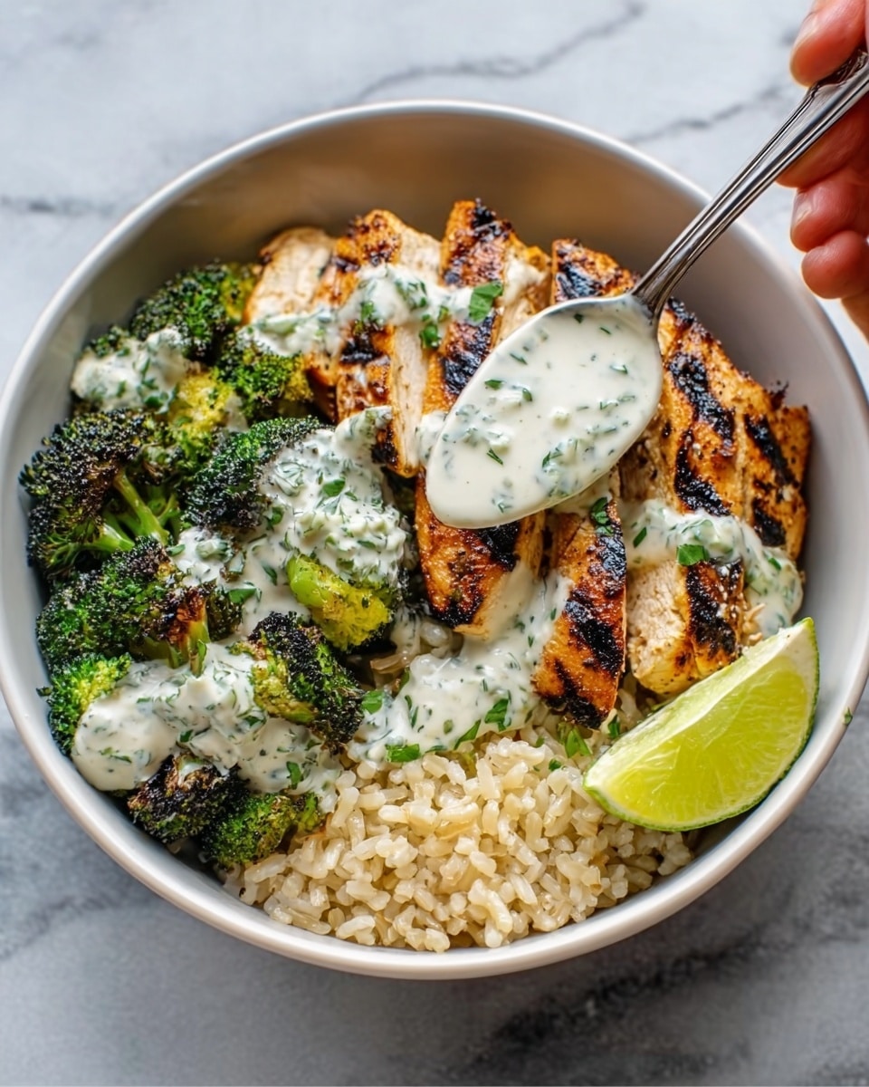 A white bowl filled with three layers of food: at the bottom, a bed of light brown cooked rice; on the right side, a lime wedge sits next to the rice; on top, three pieces of grilled chicken, showing charred grill marks and a golden brown color; the chicken is topped with white sauce speckled with green herbs; roasted broccoli florets with a dark green color and slightly crisp texture fill the left side of the bowl; a silver spoon held by a woman's hand is scooping the sauce over the chicken; the bowl sits on a white marbled surface. photo taken with an iphone --ar 4:5 --v 7