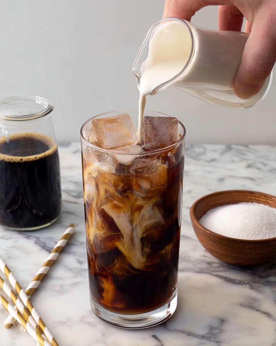 A clear tall glass filled with dark brown iced coffee and large ice cubes shows a small glass jug pouring white cream on top, creating soft swirling cream patterns inside the drink. The glass is placed on a white marbled surface alongside a brown bowl filled with white sugar, two striped white and gold paper straws, and a glass container with black coffee. A woman's hand holds the small jug pouring the cream. photo taken with an iphone --ar 4:5 --v 7