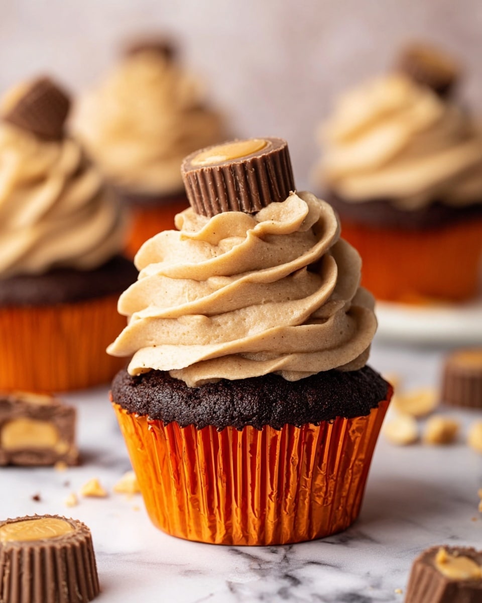 A chocolate cupcake in an orange liner sits on a white marbled surface. The cupcake has a dark, rich base topped with two large swirls of light tan frosting with smooth, creamy texture and slight darker streaks. On top of the frosting, there is a small peanut butter cup chocolate candy placed centered. In the background, there are more similar cupcakes blurred out, and pieces of peanut butter cups and peanuts scattered around. The photo taken with an iphone --ar 4:5 --v 7