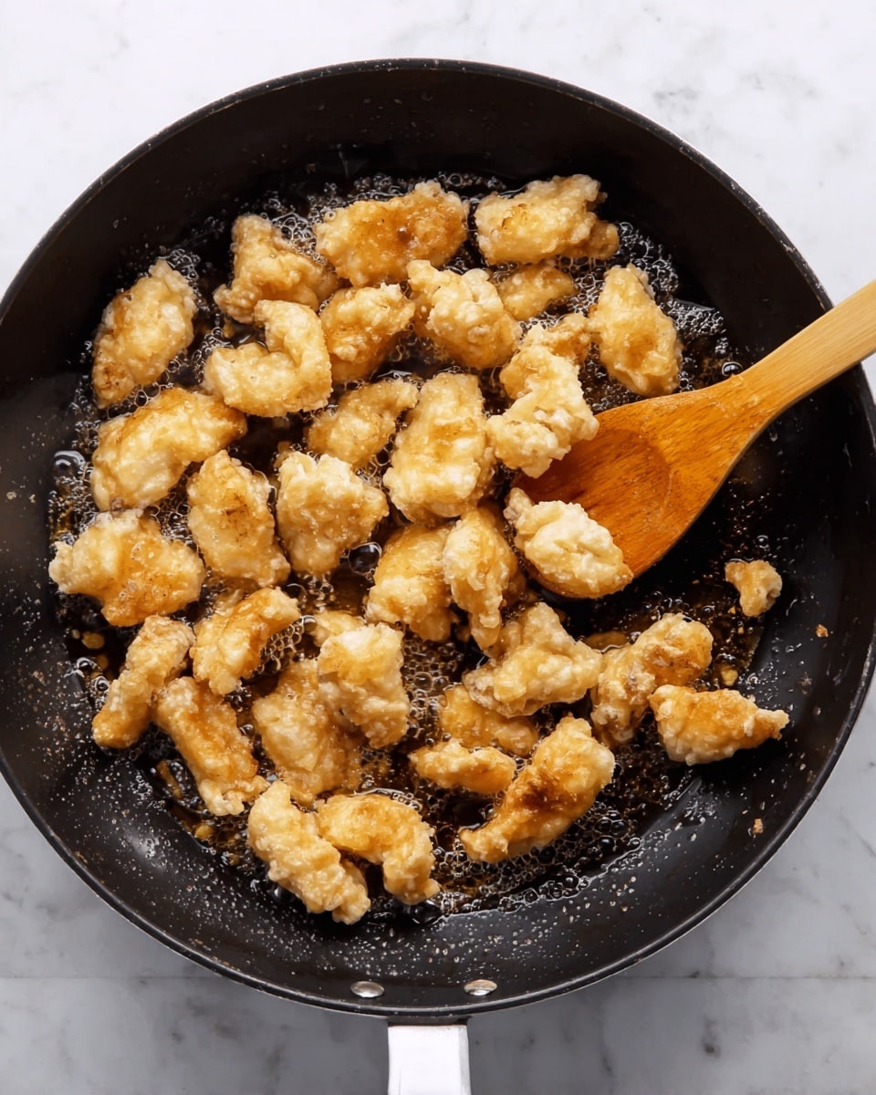 The image shows many pieces of light golden fried dough or battered food frying in a black skillet with some oil. The food pieces are irregular in shape with visible crisp, bumpy texture and slight browning around edges. A wooden spoon with a smooth texture is stirring the pieces inside the pan. The skillet has a white handle and is placed on a white marbled surface. The overall tone is warm with steam rising slightly from the hot cooking. Photo taken with an iphone --ar 4:5 --v 7