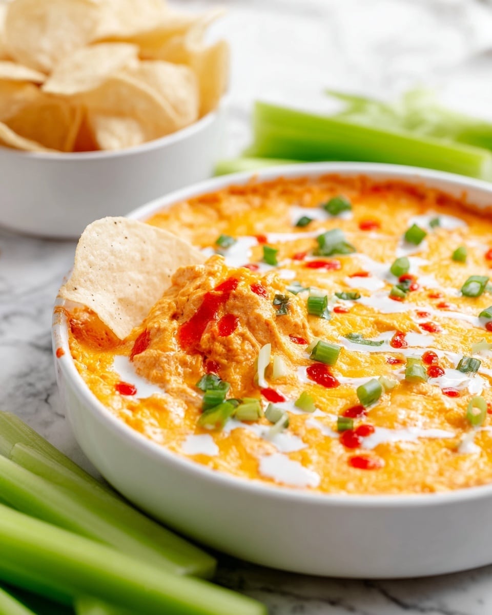 A white round dish holds a layered cheesy dip with a smooth, thick orange base topped by melted golden-yellow cheese that looks bubbly. On the surface, there are small green onion pieces scattered, bright red sauce dots, and thin white drizzles of creamy dressing. A crisp, light tan chip is partially dipped into the cheesy layer near the edge. The dish sits on a white marbled surface with fresh green celery stalks in the foreground and a white bowl filled with light tan chips slightly blurred in the background. Photo taken with an iphone --ar 4:5 --v 7