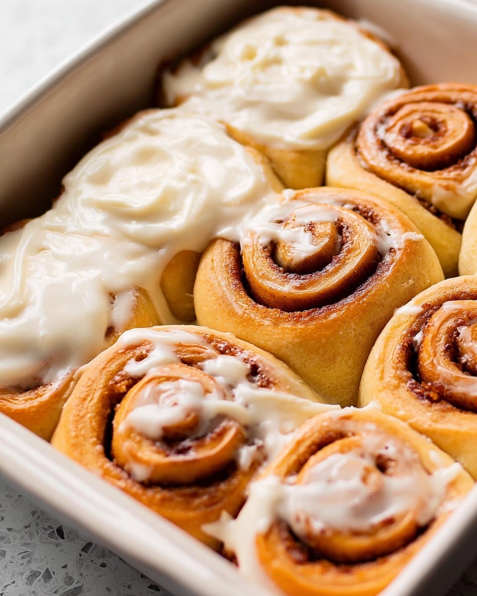 A close-up view of a white baking dish filled with seven cinnamon rolls, some covered with creamy white icing that is smooth and thick, and others without icing showing golden brown and slightly shiny spiral layers of dough with a rich cinnamon filling. The rolls are soft and fluffy, tightly packed next to each other, and the icing slightly spills over the edges of some rolls, adding texture. The dish rests on a white marbled surface. photo taken with an iphone --ar 4:5 --v 7