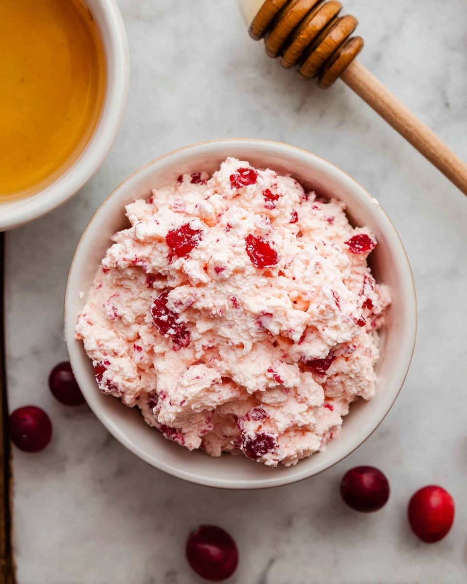 A white bowl filled with a fluffy, pale pink mixture with bright red fruit pieces scattered throughout, giving it a textured, chunky look. The bowl is placed on a white marbled surface, with a few whole red fruits around it. On the upper left, part of another white bowl with a golden liquid inside is visible, along with a wooden honey dipper handle just touching the edge of the frame. photo taken with an iphone --ar 4:5 --v 7
