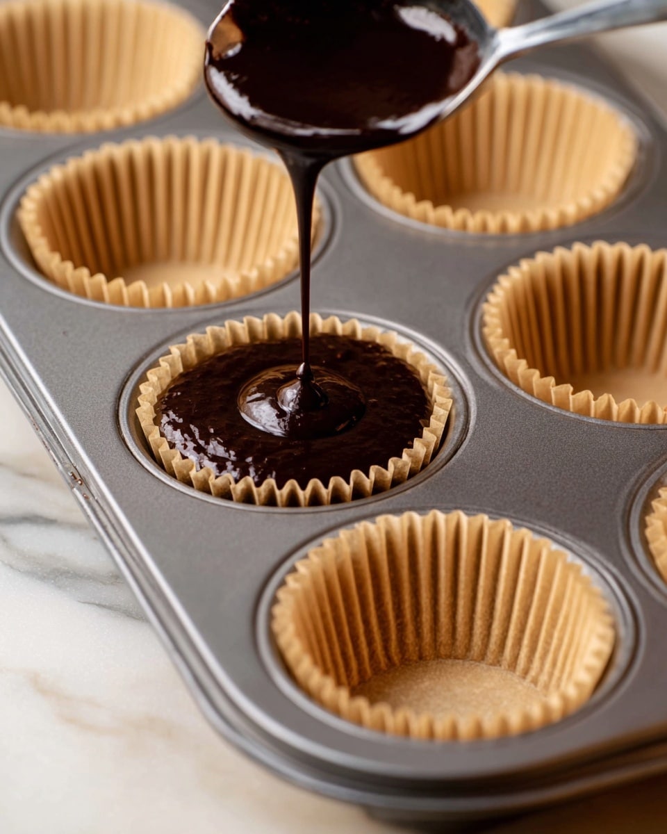 A metal cupcake tray holds six cups lined with light brown paper liners. Dark brown chocolate batter is being poured from a metal spoon into one of the liners near the center, filling it halfway. The tray rests on a white marbled surface, with the focus close on the pouring action, showing the smooth, thick texture of the chocolate batter falling into the liner below. Photo taken with an iphone --ar 4:5 --v 7