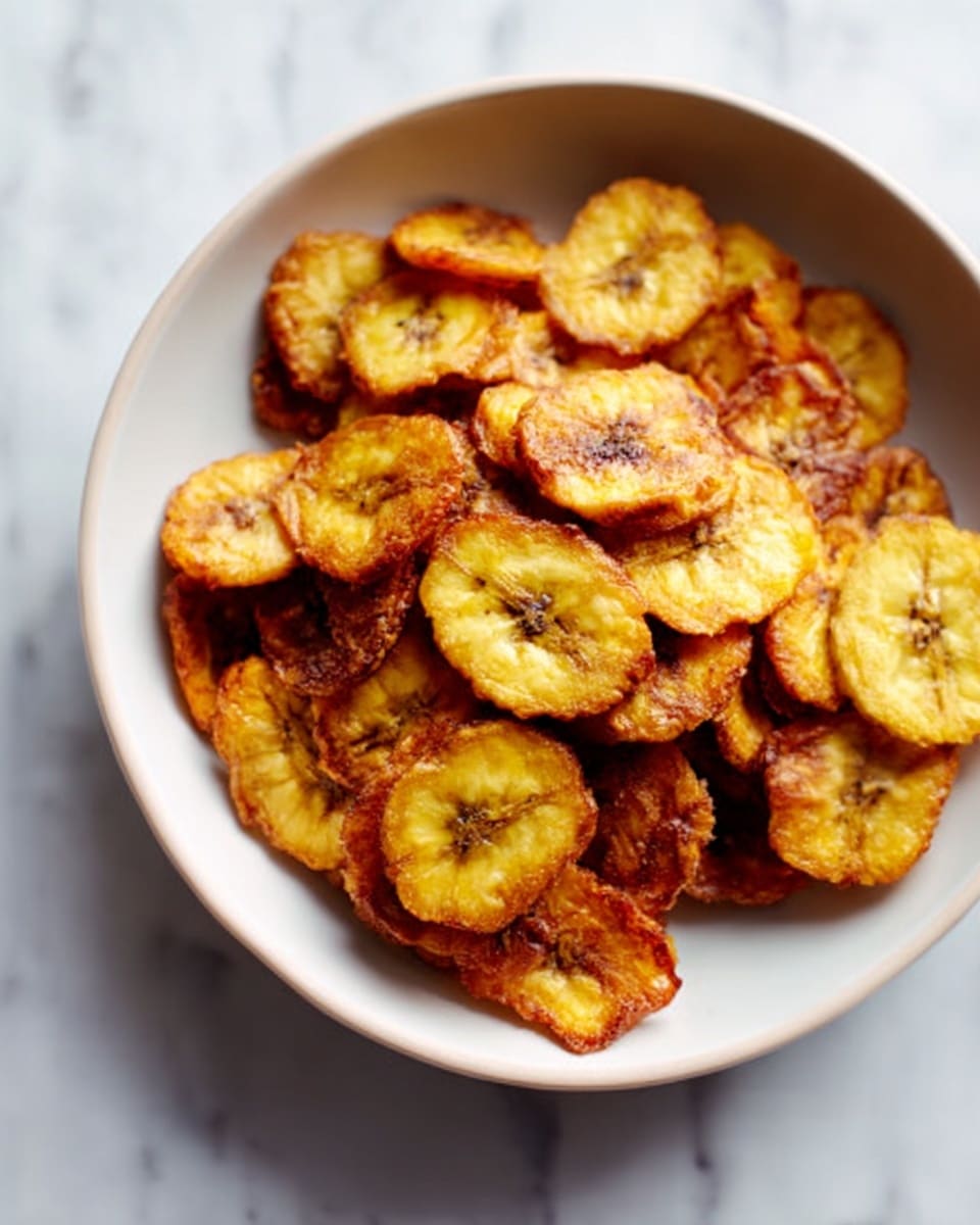 The image shows a white bowl filled with round, thinly sliced fried plantains. Each plantain slice is golden brown with darker crisply edges, showing a crunchy texture. The slices are layered in a slightly messy pile, with some overlapping and others laying flat. The bowl is set on a white marbled surface. photo taken with an iphone --ar 4:5 --v 7