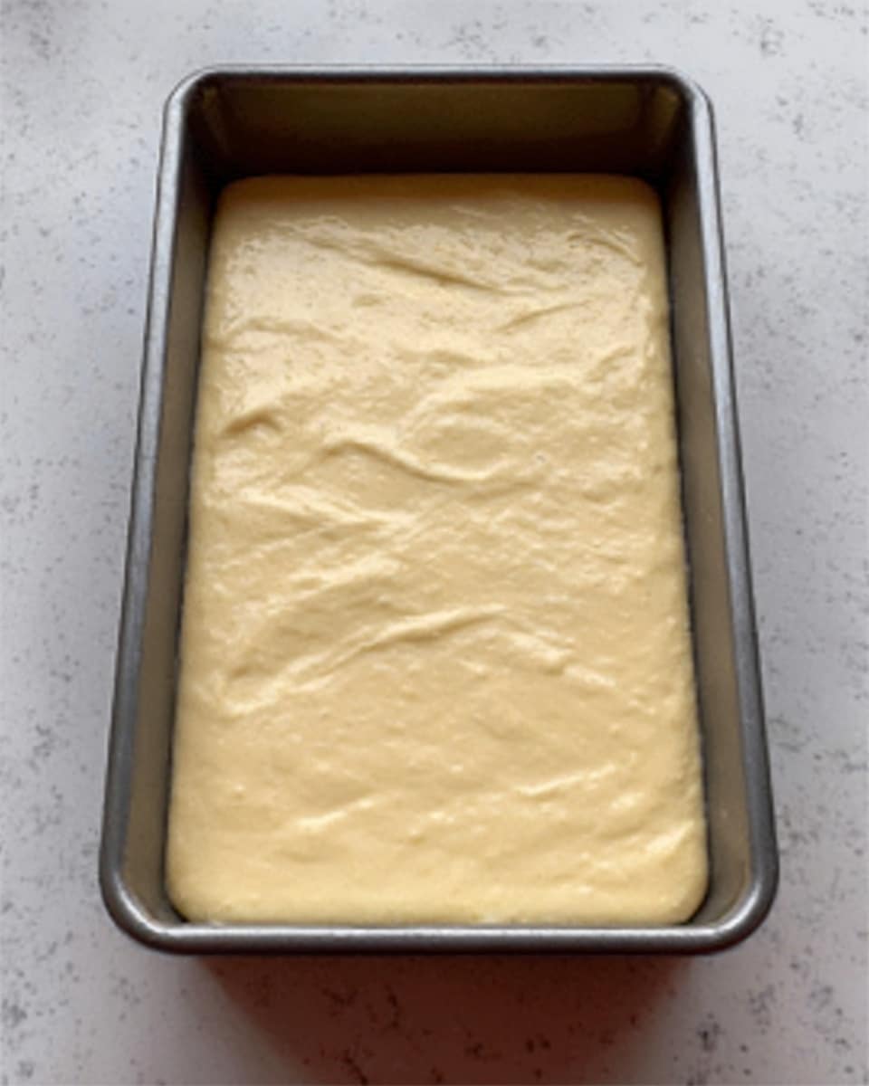 A rectangular metal tray filled with a smooth, pale yellow batter spread evenly across the surface. The batter has a slightly glossy texture with gentle swirls from spreading, and it fills the tray almost to the top. The tray is placed on a white marbled surface. Photo taken with an iphone --ar 4:5 --v 7