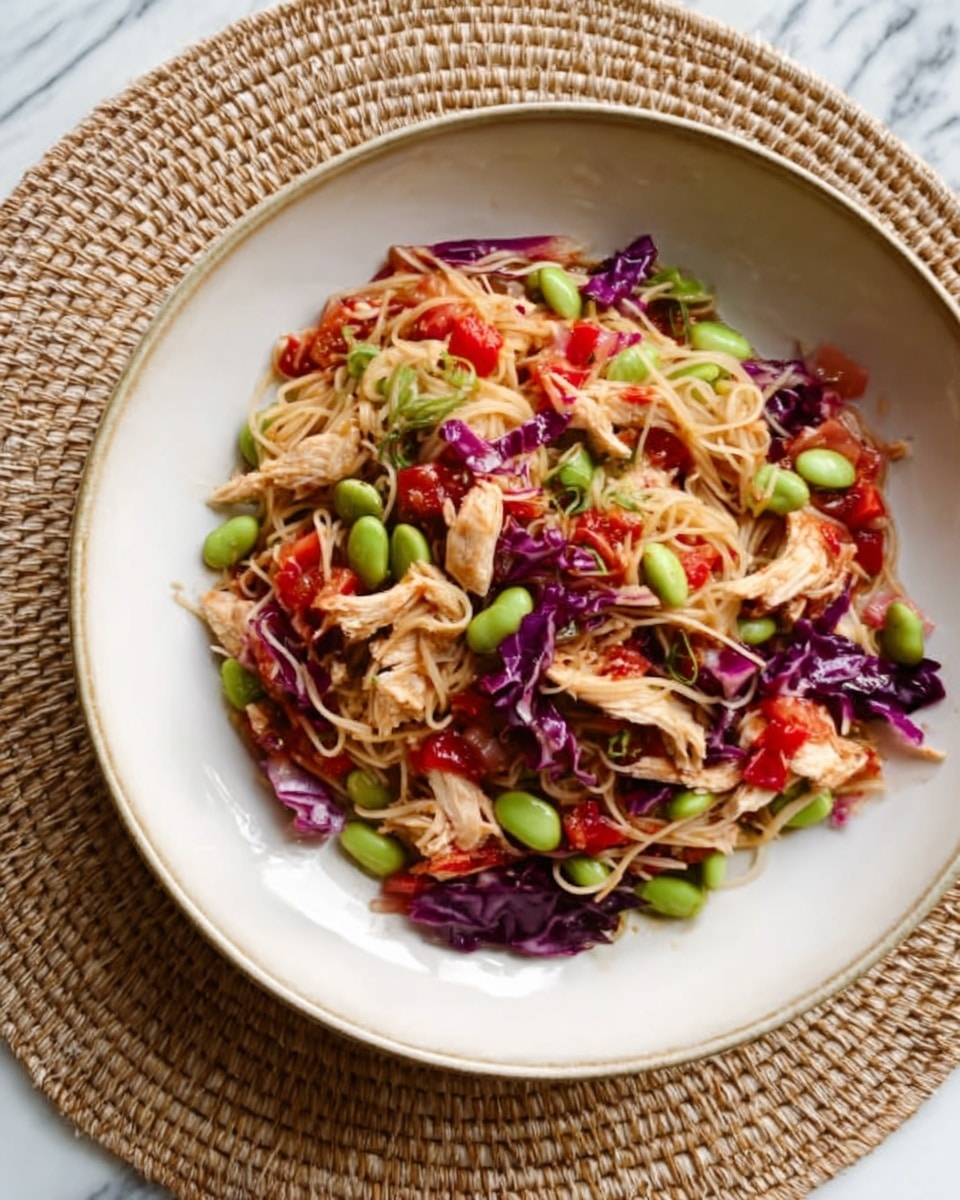 A white bowl filled with a colorful mix of thin noodles at the bottom layer, topped with shredded pieces of light-colored cooked chicken mixed with bright green edamame beans, bits of roasted red peppers, and slivers of purple cabbage. The ingredients are spread evenly through the bowl, offering a variety of textures and colors with a light sauce coating them. The bowl sits on a round woven placemat on a white marbled surface. Photo taken with an iphone --ar 4:5 --v 7
