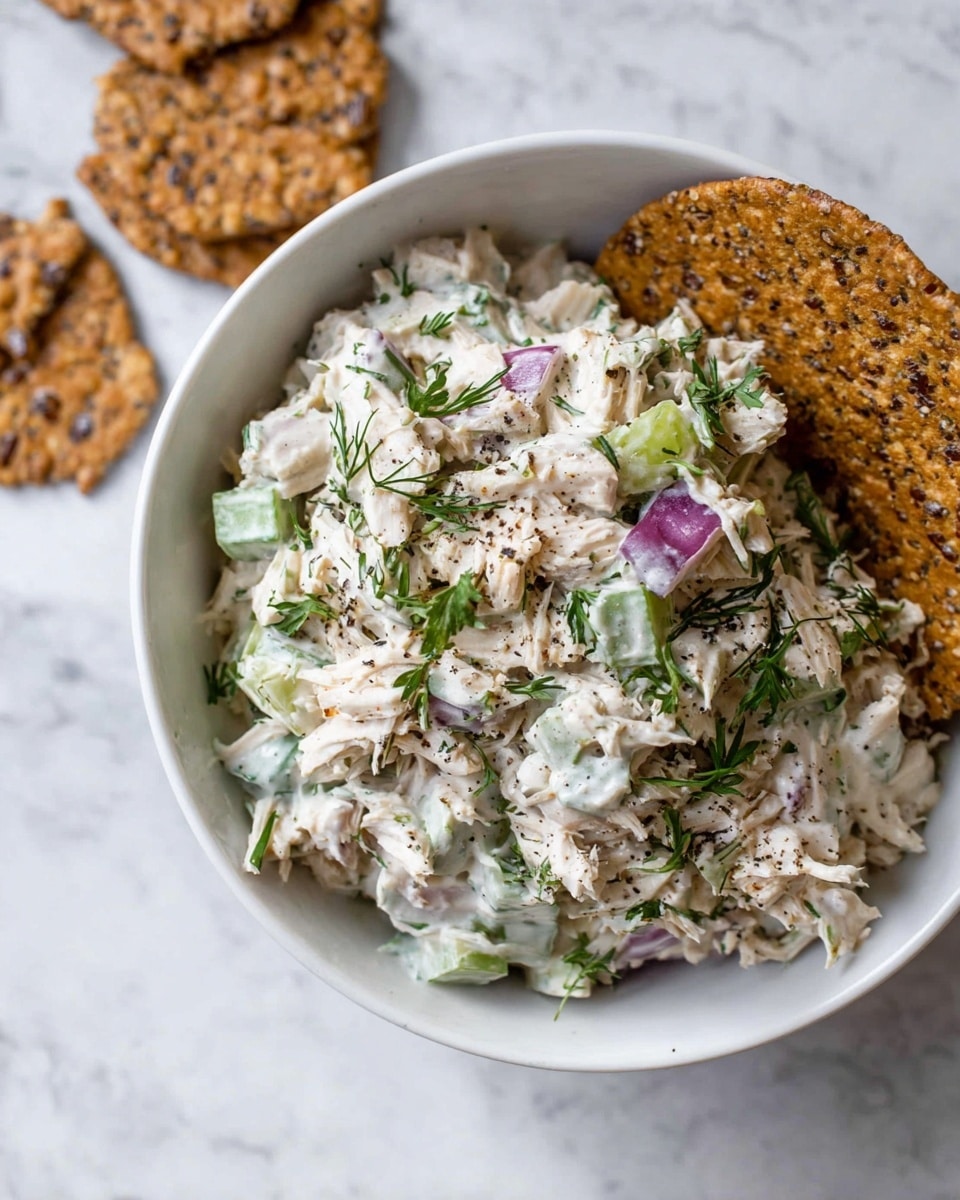 A white bowl filled with a creamy salad that has several layers of shredded white meat mixed with chopped green celery and pieces of purple onion, all coated in a thick white dressing. The salad is sprinkled with fresh green herbs and ground black pepper. A round, dark brown cracker with visible nuts and seeds is placed on the side of the salad inside the bowl. The bowl sits on a white marbled surface with more of the same dark brown crackers scattered nearby. Photo taken with an iphone --ar 4:5 --v 7