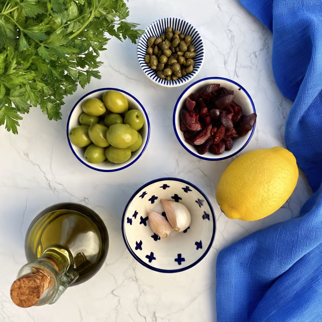 On a white marbled surface, there is a bunch of fresh green parsley in the top left corner. Below it, a small white bowl with blue trim holds whole green olives. To the right, another small white bowl with blue trim contains dark purple olives cut in halves or pieces. Above those olives, there is a small white bowl with blue stripes filled with whole capers. Near the center bottom, a small white bowl with navy blue crosses holds two garlic cloves. A yellow lemon is placed to the right of the bowls near the center. At the bottom left, there is a round glass bottle filled with olive oil and sealed with a cork. A blue cloth is spread out on the right side of the image. Photo taken with an iphone --ar 4:5 --v 7