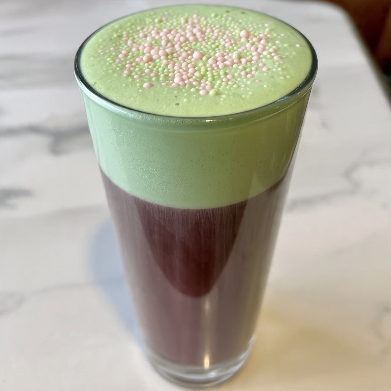 A tall clear glass filled with two layers, the bottom layer is dark brown liquid with a smooth texture, and the top layer is thick light green foam, covering the whole surface of the glass. Small round sprinkles in pale pink and light green are scattered evenly over the foam. The glass is set on a white marbled surface. Photo taken with an iphone --ar 4:5 --v 7