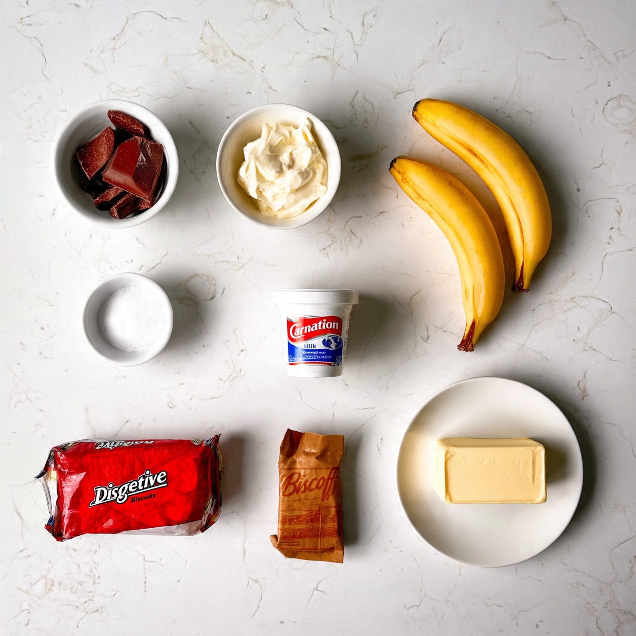 The image shows ingredients for a recipe arranged neatly on a white marbled surface. There are two yellow bananas at the top right, a small white bowl with a few pieces of dark brown chocolate on the top left, and next to it is a small white bowl filled with cream. Below that is a small white bowl with white powder, possibly salt. In the center is a white container of Carnation evaporated milk. Toward the bottom left, there is a package of red Digestive biscuits, next to it a brown bag of Biscoff spread, and on the right side of this row is a white plate with a block of light yellow butter. Photo taken with an iphone --ar 4:5 --v 7